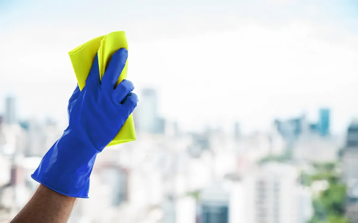 Mano con guante azul limpiando un ventanal con paño amarillo frente a la ciudad